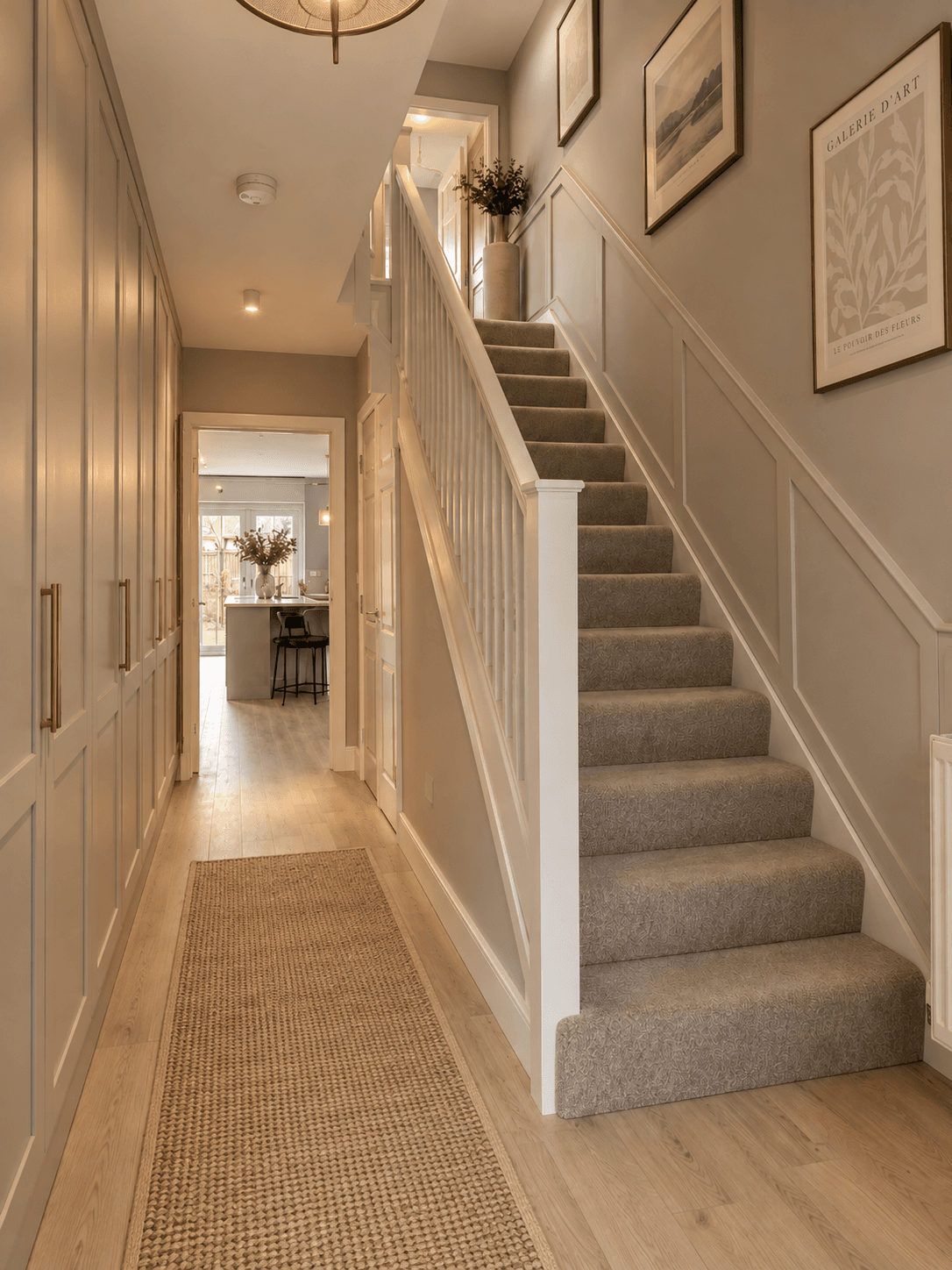 Modern new build hallway with full height built in storage, warm greige walls, wall moulding, neutral carpeted stairs, oak flooring and soft layered lighting.